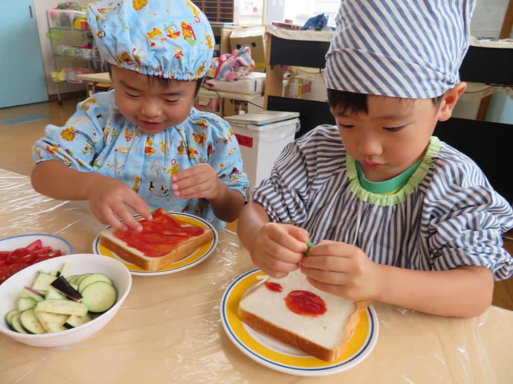 ４月から園庭でキリン組さんが育てている夏野菜を使って、夏野菜ピザを作りました♪
作り方は、とっても簡単☆
①パンにケチャップをぬる
②野菜をのせる
③チーズをのせる
④焼いて出来上がり♪

真っ白なパンに思い思いの想像膨らませながら・・・
「森みたいなピザにする」「地図みたいにする！」と盛り上がっていました！！
