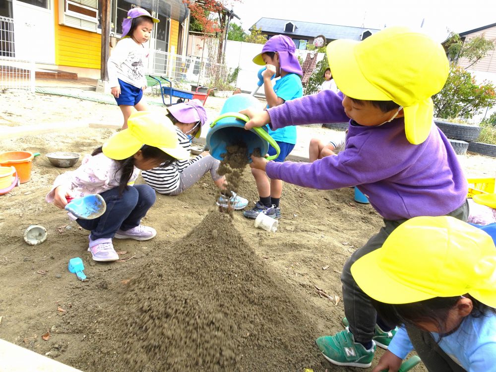 隣で大きな砂山を作るキリン組さんを見て、
「キリン組さんより大きな山を作りたい！」と
スコップやバケツを持って集まったゾウ組さん

「もっと土をかけんと！」とやる気満々のKくん
バケツいっぱいに土を入れて、どんどんかけていきます！