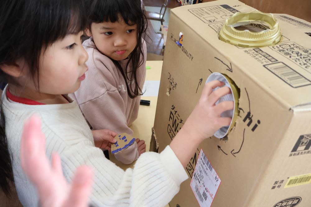 段ボールに穴をあけ
取っ手にちょうどいい廃材も発見！
まわす方向の矢印も書いてばっちり！
 順調♡
・ ・ ・ のはずが ・ ・ ・ 
「なんかすぐ取れる！」

まわすと取れてしまう取っ手に困った様子 ・ ・ ・

「段ボールとくっつけたらまわらないし・・・」
「まわす人に気をつけてもらったらいいかな・・・」
と言いつつも、やっぱり取れることが気になる

３人で時間をかけてなぜとれてしまうの考える！

「このすき間があいてるからダメなんじゃない？」
「すき間を埋めてみよう⭐︎」

すき間に新聞紙を入れてふさぐと
ぴったりフィット♡
