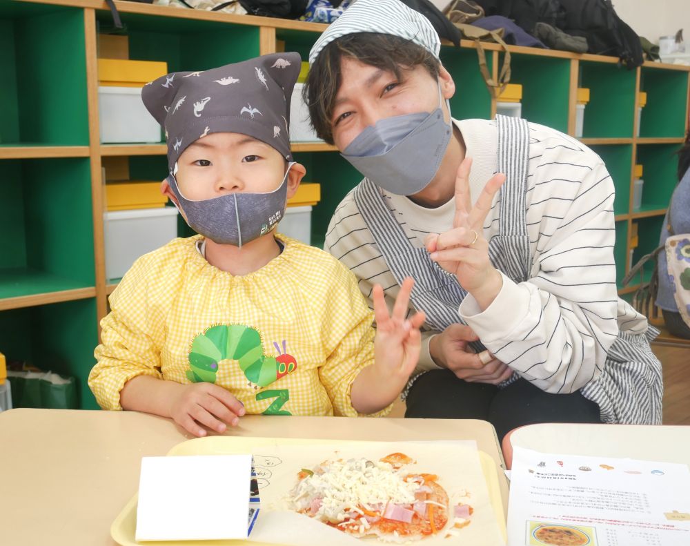 野菜の次は、ベーコンをのせて、仕上げはピザに欠かせないチーズを全体に散らします。
「もっとチーズのせたいなぁ。おうちでも今度しようね！チーズ多めね！」
「ご飯のピザ、どんな味かなぁ。シーチキン入れてもおいしそうじゃね♪」
と、家庭でのクッキングにつながりそうな、嬉しい声も聞こえてきました♡

トッピングが終わったら・・・
頑張った記念に写真をパチリ♪
お父さんの嬉しそうな笑顔♡

色とりどりのみんなのライスピザ、さっそく給食室で焼きましょう！！