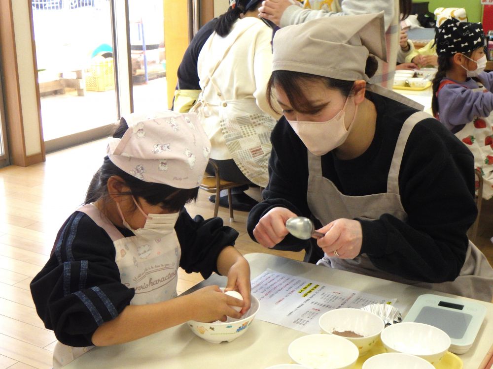 グループの材料が集まったら、今度は一人分ずつに分ける作業。
はかりを使い、必要な量を取り分けます。
親子で顔を寄せ合い計量する姿がほほえましたったです♡
「こぼれないように気をつけようね」と、慎重に、少しずつ量ります。
慣れない道具でも、おうちの人となら楽しんで使いこなしていますね♪

材料がそろったら、いよいよ生地作りがスタート！
卵を割る作業が、ちょっとドキドキの子ども達。
そんなときは、お母さんのアドバイスを聞いて♡
「そしたらそこに親指を入れて・・・」と、同じポーズで教えてくれます。
「えいっ」と勇気を出して、、、おうちの人と頑張る時間も楽しいね♪