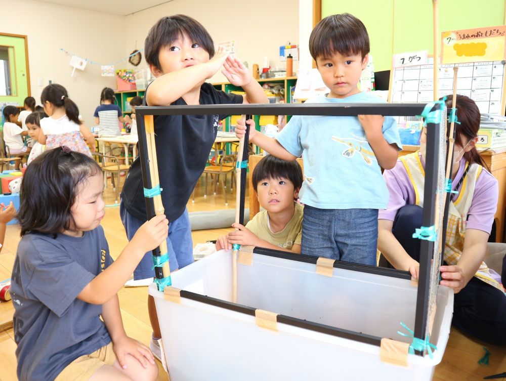 “おうちの囲いをどうする・・？”　とみんなで話していると・・
「こんな屋根みたいのとか？」
「四角いお家もいいと思う！」
「壁とか網みたいなのつけんと、逃げるよね」
「大きい木も入れてあげたら喜びそう！」」
色々なアイディアが・・♡
　
みんなで柱を支えたり、紐をくっくたりしながら
カブトムシのお家づくりが進んでいく・・
